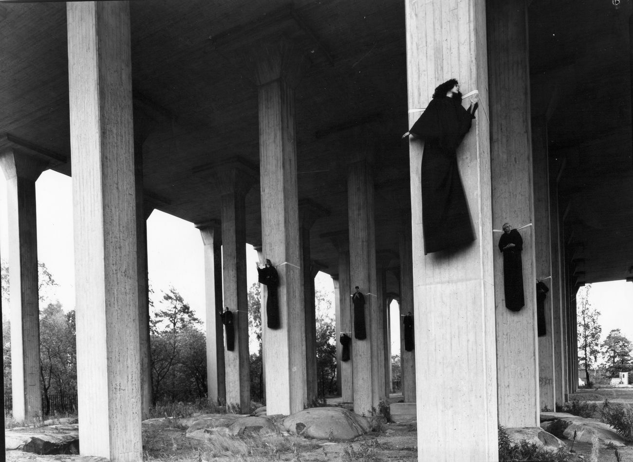 Svartklädda figurer fastspända högt uppe på betongpelare under en viadukt.